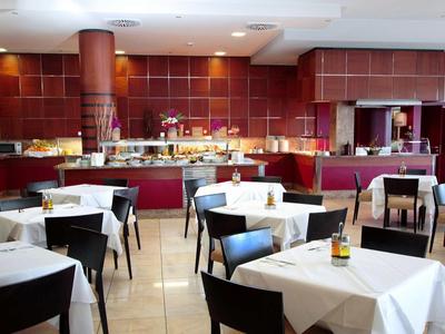 Restaurant mit quadratischen Tischen, weißen Tischtüchern und dunklen Stühlen vor roter Wand.