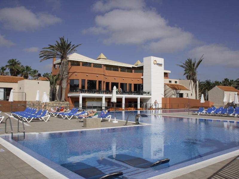 Modern hotelcomplex met groot buitenzwembad en ligstoelen onder een zonnige hemel.