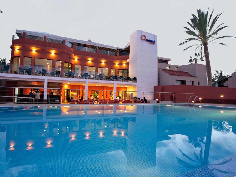 Verlicht hotel met buitenzwembad en palmbomen bij zonsondergang.