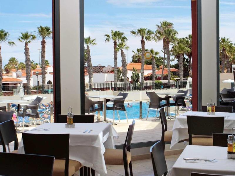 Restaurant met gedekte tafels en uitzicht op buitenzwembad en palmbomen.