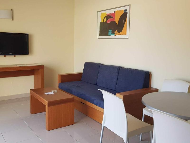 Eenvoudig ingerichte woonkamer met blauwe bank, tv, kleine tafel en eettafel met witte stoelen.