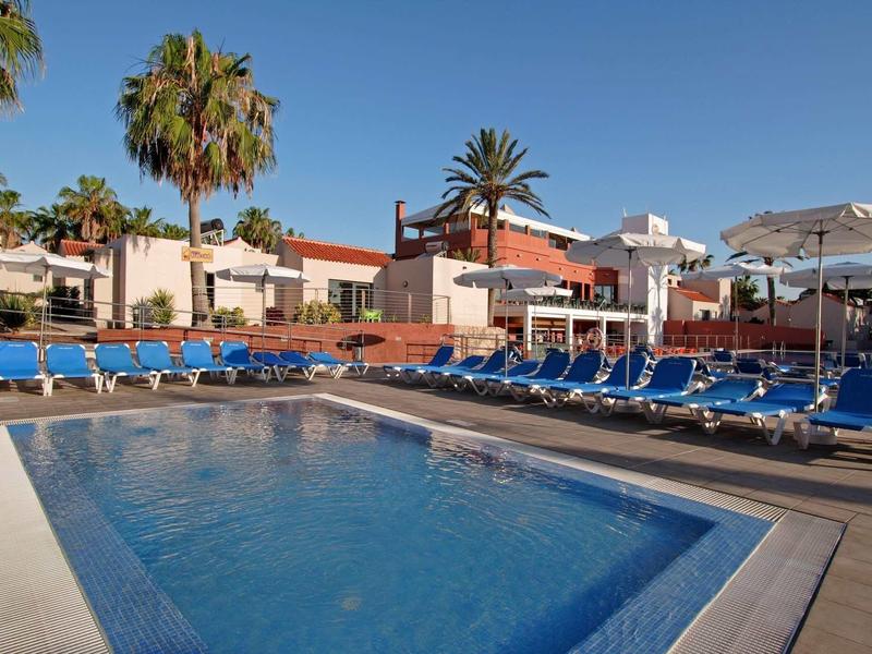 Heldere buitenzwembad met blauwe ligstoelen en parasols naast hotelgebouwen en palmbomen.