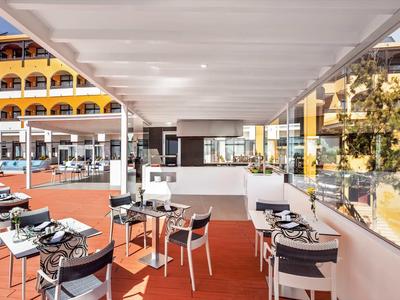 Moderner Außenbereich eines Restaurants mit Tischen und Stühlen auf rotem Bodenbelag und weißem Dach.
