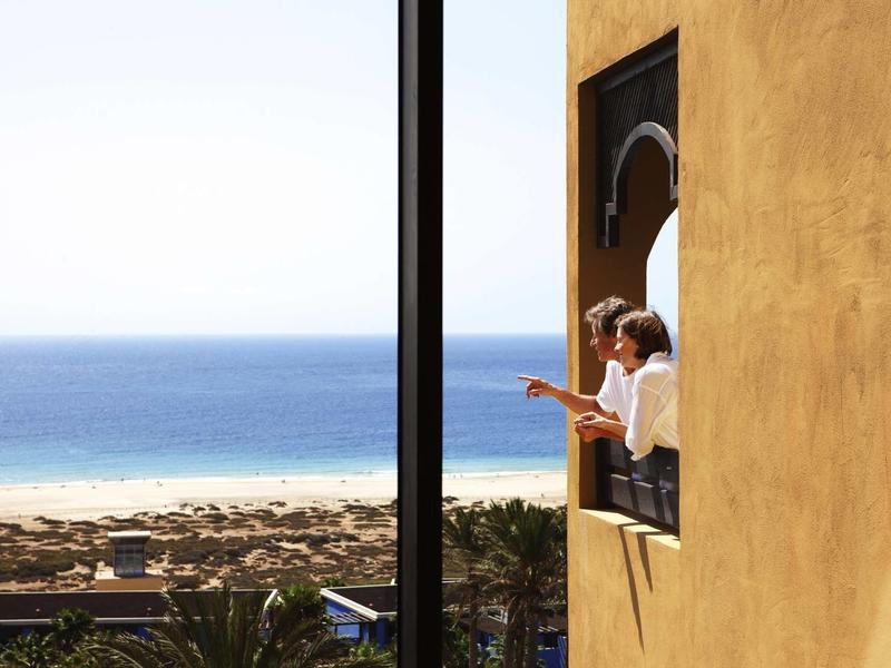 Zwei Personen stehen am Fenster eines gelben Hauses mit Blick auf Strand und blauen Ozean.