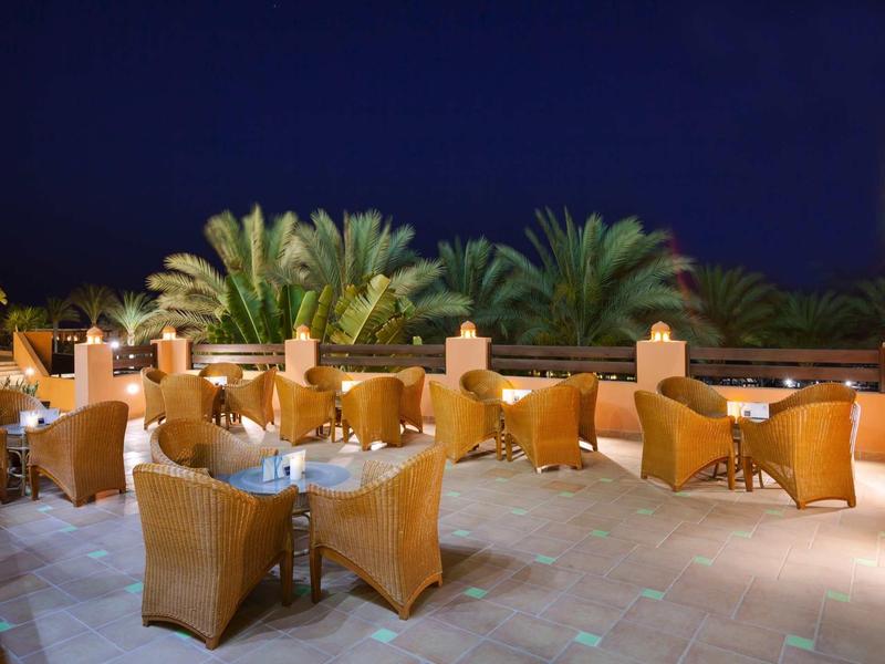 Freiluft-Café mit braunen Korbsesseln und Tischen, Palmen im Hintergrund bei Nacht.