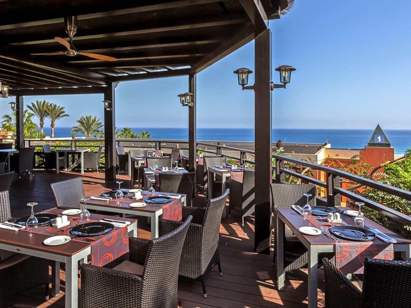 Offene Terrasse mit gedeckten Tischen, Blick aufs Meer, Palmen und blauer Himmel im Hintergrund.