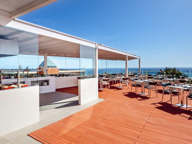 Offene Terrasse mit Holzboden, Tischen, Stühlen und Blick auf das Meer bei klarem Himmel.