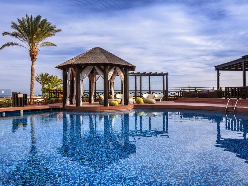 Großer blauer Pool mit Pavillon, Palmen und offenem Himmel im Hintergrund.