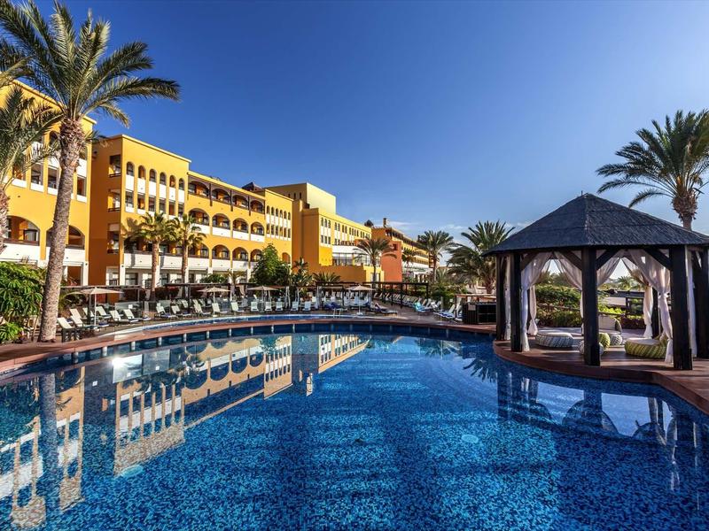 Großer Pool mit Pavillon, umgeben von Palmen und gelbem Hotelgebäude unter blauem Himmel.