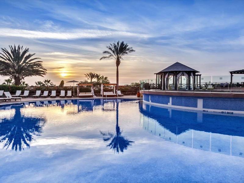 Großer Pool mit blauer Wasserfläche, Palmen und Sonnenuntergang am Horizont, gemütliche Lounge-Bereiche.
