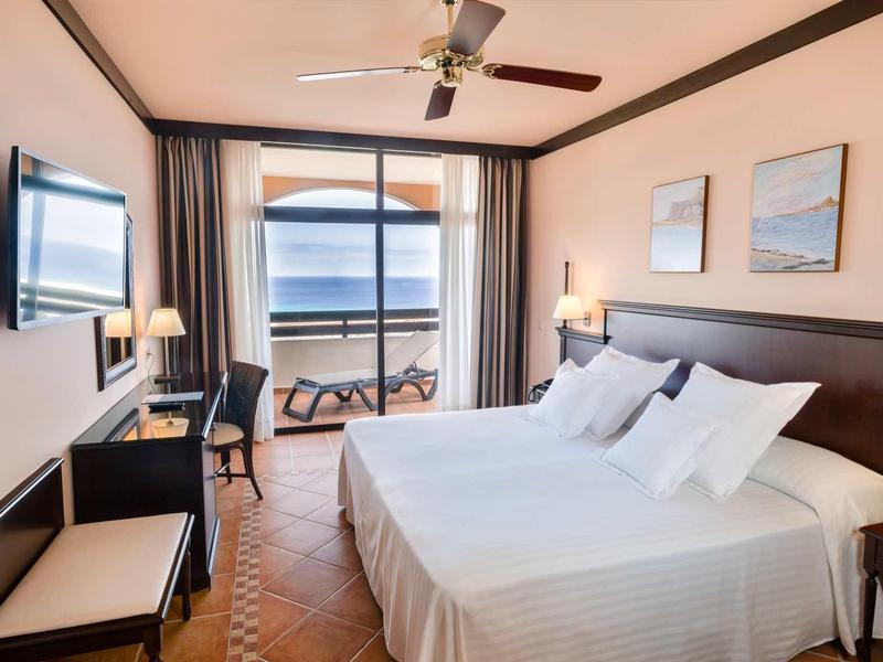 Modernes Hotelzimmer mit Doppelbett, Deckenventilator, Schreibtisch und Meerblick auf dem Balkon.