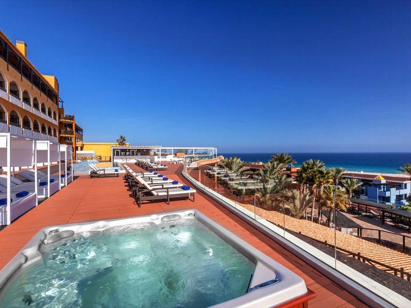 Rooftop mit Whirlpool, Liegen und Blick auf Palmen, Strand und blaues Meer unter klarem Himmel.