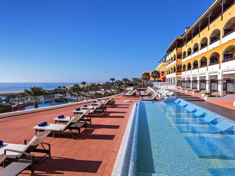 Langgezogenes Schwimmbecken mit blauer Wasseroberfläche neben Hotel mit gelber Fassade und Sonnenliegen.