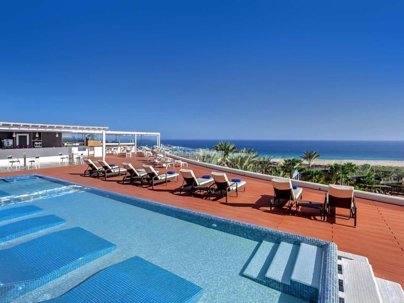 Moderner Pool mit Liegestühlen, rotem Boden und Meerblick unter klarem blauem Himmel.