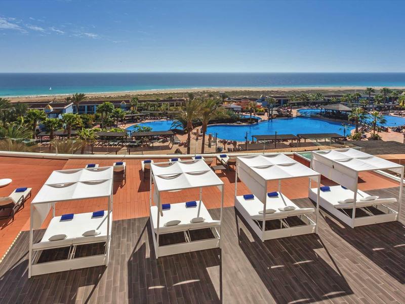 Luxuriöse Sonnenterrasse mit weißen Liegen, blauem Pool und Meer im Hintergrund unter klarem Himmel.