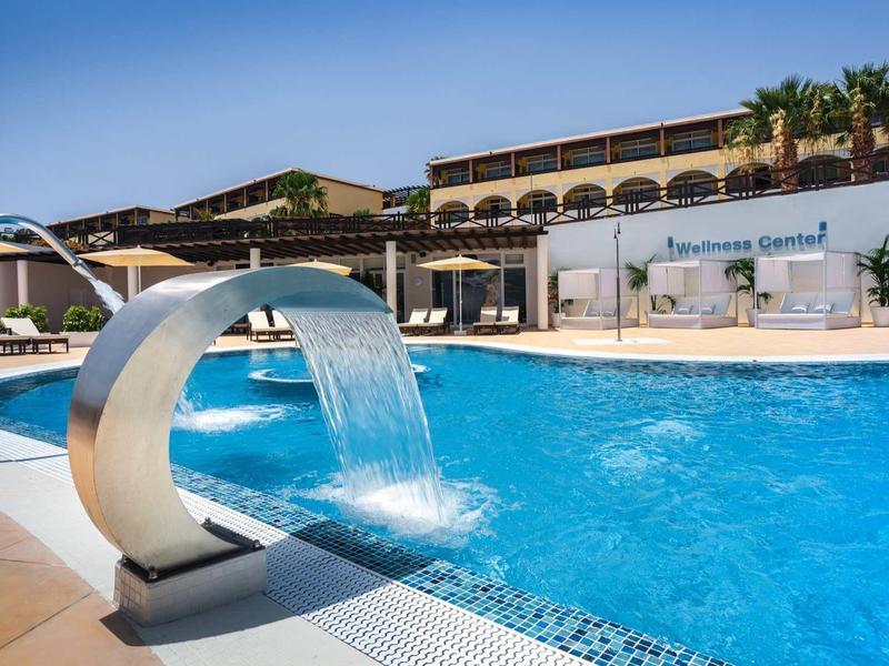 Großer Außenpool mit Wasserfall, umliegenden Liegestühlen und modernem Hotelgebäude im Hintergrund.