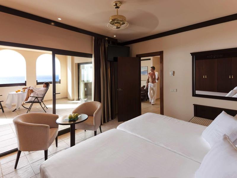 Helles Schlafzimmer mit weißem Bett und zwei Sesseln, offen zu Terrasse mit Meerblick und Balkonmöbeln.