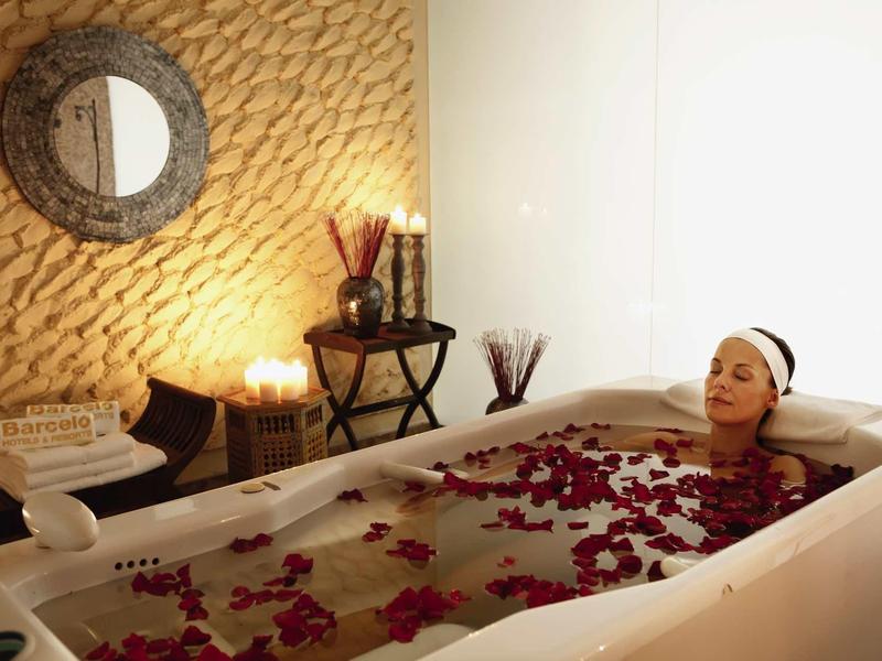 Frau entspannt in Badewanne mit Rosenblättern, warme Beleuchtung und rustikale Wand im Spa-Ambiente.