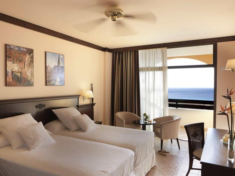 Hell eingerichtetes Hotelzimmer mit zwei Einzelbetten und Blick auf das Meer durch große Fenster.