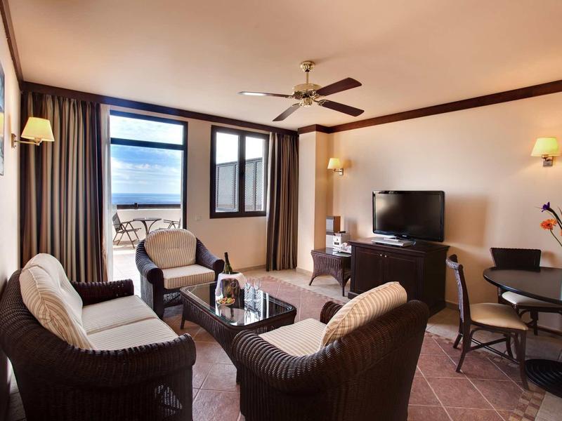 Gemütliches Wohnzimmer mit braunen Rattanmöbeln, Glastisch, TV und großem Fenster mit Vorhängen.