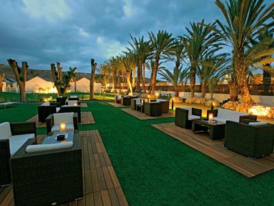Außenlounge mit Palm Bäumen, modernen Sitzgruppen, Holzterrasse und grünem Kunstrasen bei Abenddämmerung.