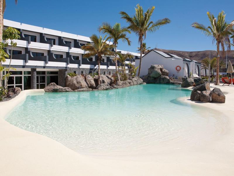 Hotel met groot zwembad, palmbomen en wit zand onder een heldere blauwe lucht.