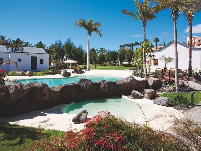 Hotel tuin met zwembad, palmbomen en ligstoelen onder een blauwe lucht