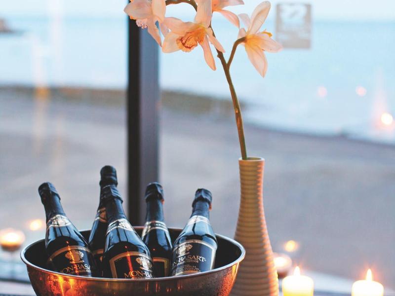 Tafel met champagneflessen, glazen en een vaas met bloemen bij een raam met uitzicht op zee