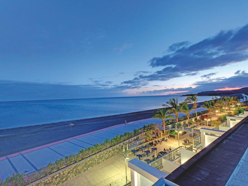 Avondzicht op een verlicht strandrestaurant met palmbomen naast rustige zee en blauwe lucht.