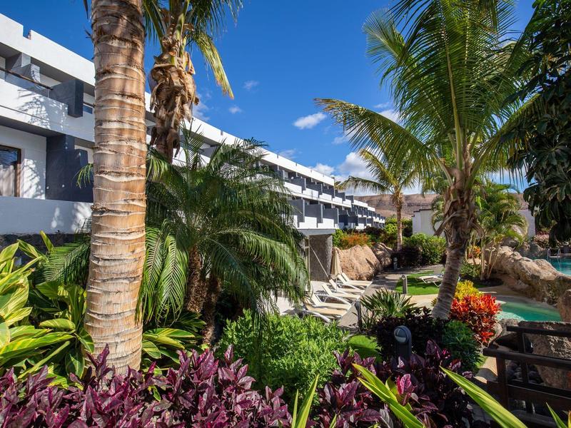 Modern meerlaagse hotel omgeven door palmbomen en weelderige tropische planten bij een heldere lucht.