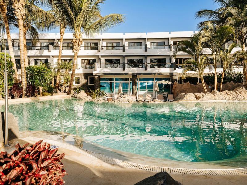 Modern hotel met groot zwembad, palmbomen en heldere blauwe lucht op een vakantiebestemming.