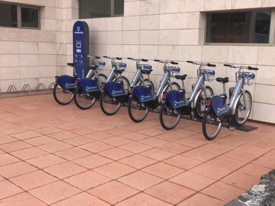 Sei biciclette blu a noleggio allineate su pavimento piastrellato rosso davanti a un muro bianco.