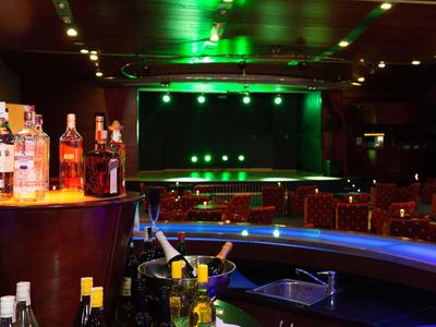 Interno di un bar con bottiglie, bancone illuminato e palco sullo sfondo con luci verdi.