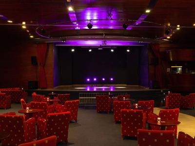 Sala teatrale vuota con sedute rosse e palco illuminato da luce viola.