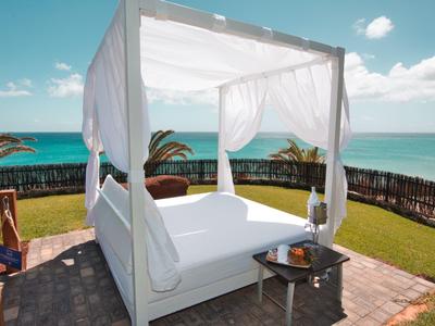 Letto a baldacchino bianco su terrazza con vista sul mare e giardino verde in giornata soleggiata.
