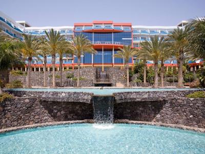 Hotel con cascata e palme davanti a un edificio moderno sotto un cielo sereno.