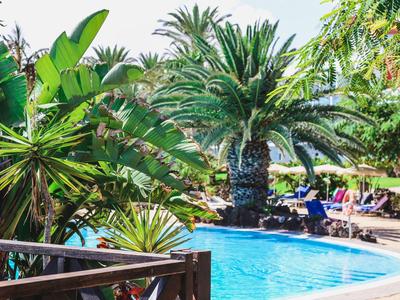 Piscina circondata da piante tropicali e palme, con lettini e ombrelloni sullo sfondo.