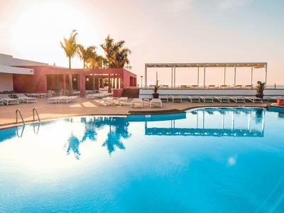 Grande piscine d'hôtel au coucher du soleil avec palmiers et espaces de détente au bord de la mer.
