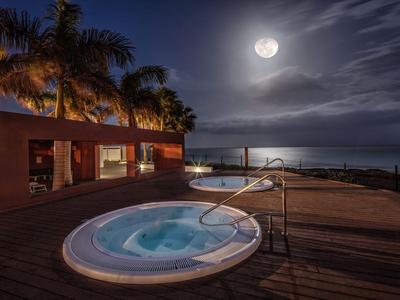 Lit jacuzzi on wooden deck overlooking the sea under a full moon at night.