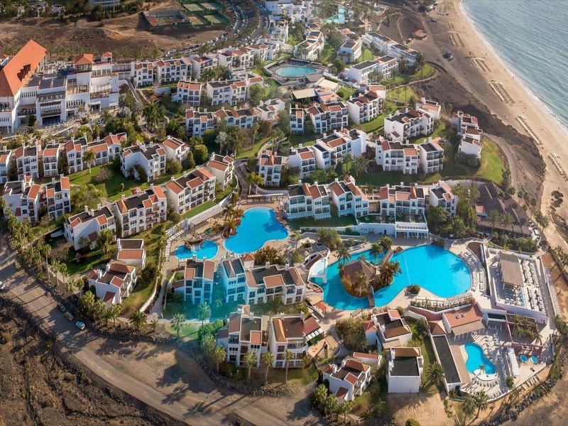Vue aérienne d'un grand complexe balnéaire avec plusieurs piscines et jardins verts au bord de la mer