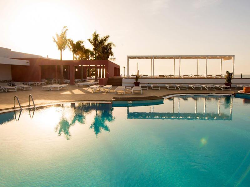 Grande piscine extérieure avec chaises longues et palmiers au coucher du soleil.