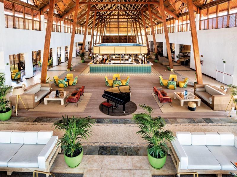 Grand salon d'hôtel lumineux avec plafond en bois, canapés et plantes.