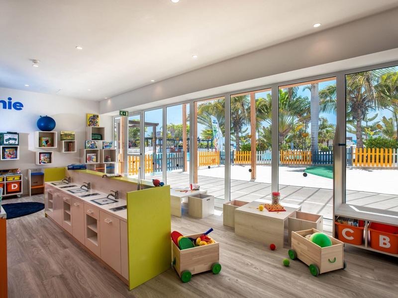 Salle de jeux lumineuse pour enfants avec des jouets, de grandes fenêtres et vue sur la piscine et les palmiers.