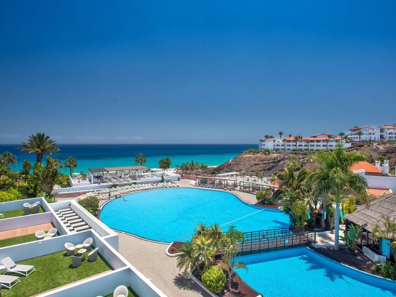 Complex hôtelier avec deux piscines, des palmiers et vue sur la mer bleue sous un ciel ensoleillé.