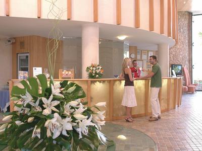 Helle Hotelrezeption mit Blumenarrangement, zwei Personen am Empfang und großem Eingangsbereich.