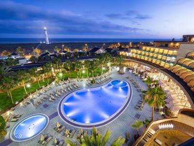 Großer beleuchteter Pool und Hotel mit Balkonzimmern bei Nacht, dahinter Meer und Leuchtturm.