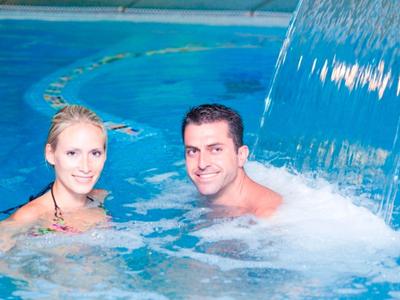 Zwei Personen entspannen im blauen Wasser neben einem Wasserfall im Schwimmbad.