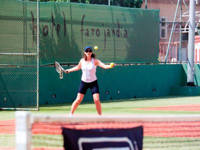 Frau spielt Tennis auf grünem Tennisplatz vor grünem Gitterzaun und rotem Gebäude im Hintergrund.