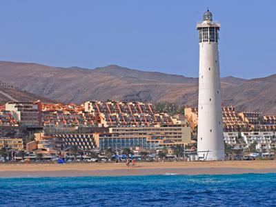 Hoher weißer Leuchtturm am Strand vor Bergen und Häusern unter klarem blauem Himmel.