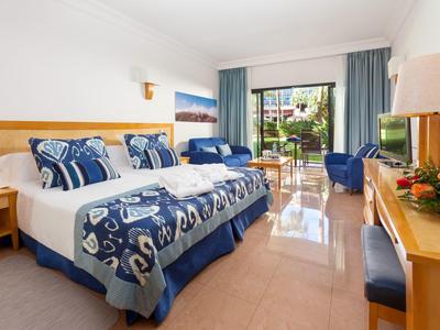 Helles Hotelzimmer mit Doppelbett, blauen Vorhängen, Sitzbereich und Gartenterrasse.
